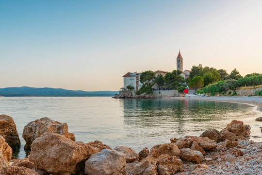 Rocky pebble beach with calm Adriatic water, historic seaside church and Mediterranean architecture in Bol town at sunset on Brac Island, Croatia. European travel destination for island tourism