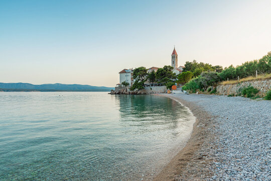 Pebble Martinica beach with crystal clear Adriatic water, historic coastal church and Mediterranean architecture in Bol town on Brac Island, Dalmatia, Croatia. Travel destination, island tourism