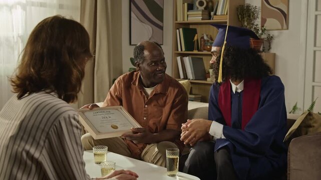 Handheld shot of Black middle aged father showing graduation certificate to Caucasian mother and young son in cap and gown, then clinking glasses. Ideal for college and family milestone campaigns