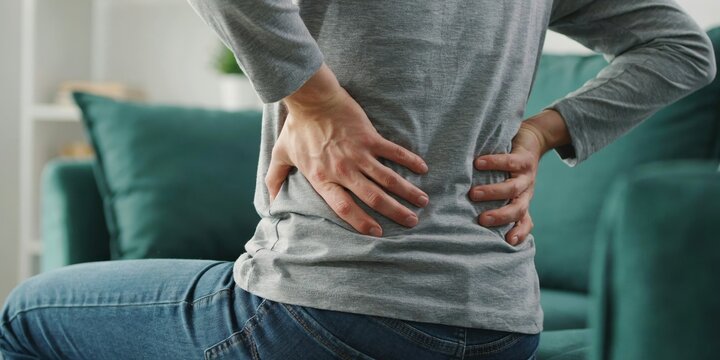 Person suffering from back pain holding hands on lower back sitting on sofa at home.