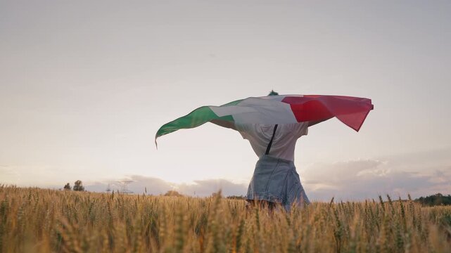 Woman walks through a golden wheat field carrying the Italian flag toward the setting sun. Symbol of patriotism, freedom, rural heritage, agriculture and pride in Italy.