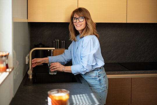 Smiling mature woman filling a glass of water from modern kitchen tap