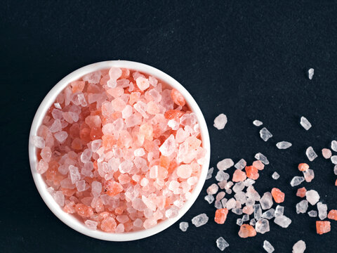 Coarse pink himalayan salt in a small white ceramic bowl on black slate background. High angle top view flat lay composition with sharp details and copy space. Perfect for cooking or wellness