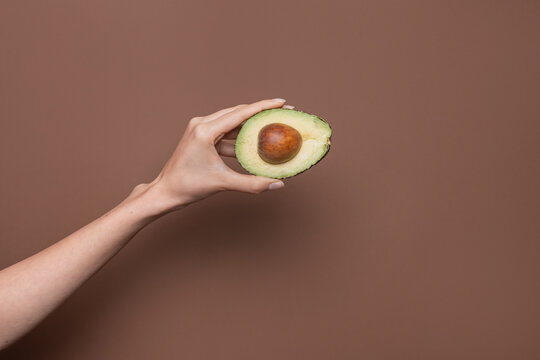 Hand holding half avocado with pit on brown background