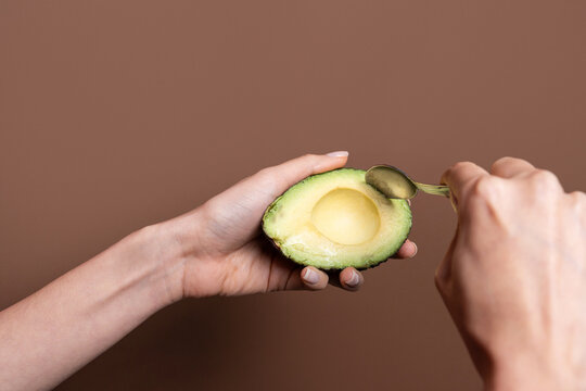 Hand scooping avocado half with spoon on brown background
