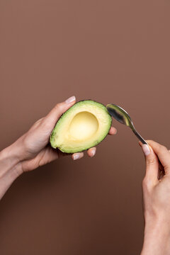 Hands holding half avocado and spoon on brown background