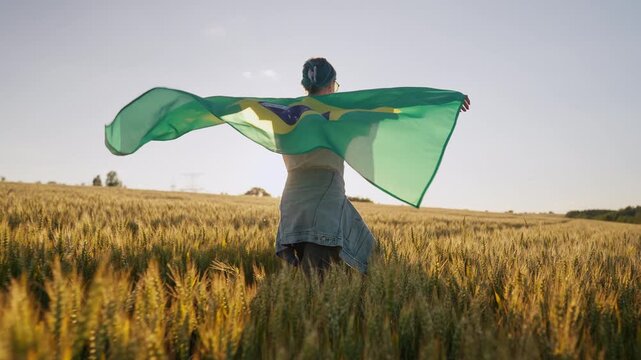 Excited Brazilian woman walking across golden wheat field, holding national flag as it flutters behind. National pride, celebration, connection to football culture in South America.