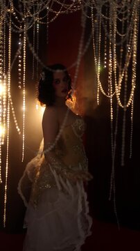 Female burlesque performer lifts voluminous tulle skirt beside dense hanging crystal bead strands, showing fringe, sequins, gloves, and dramatic warm light in dim stage setting, with burlesque