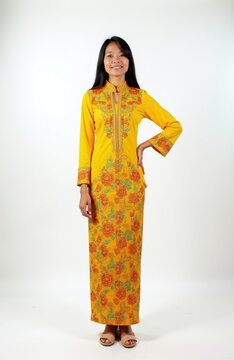 Young South east Asian woman wears traditional yellow baju kurung dress with floral embroidery. Smiling female model stands on plain white background. Full length studio portrait of person in ethnic
