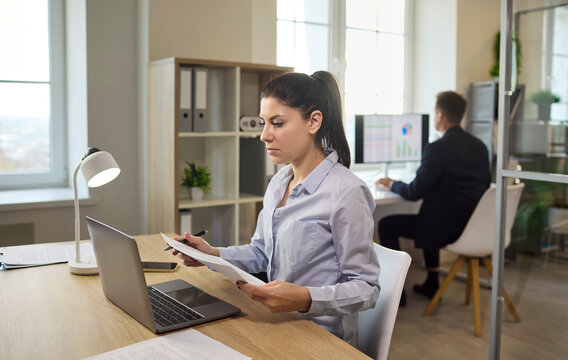 Woman analyst in office with laptop. She reviews paperwork on a computer, preparing a finance report in a modern workspace with light. Clear concept of business finance analysis at work.
