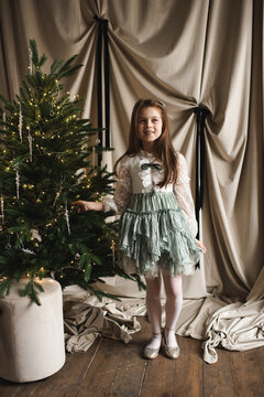 Smiling child girl 5-6 year old wearing princess dress decorating Christmas tree over beige background. Winter holiday season.