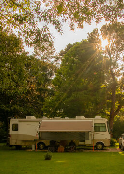 An RV is parked on a grassy patch surrounded by tall trees. Sunlight filters through the branches, creating a warm, peaceful atmosphere.