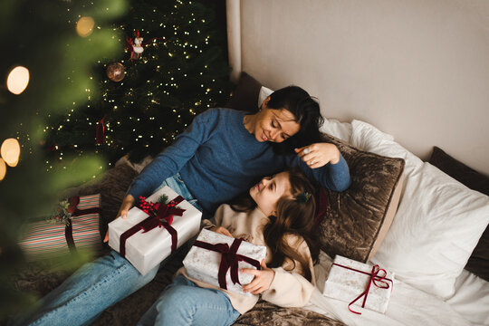 Happy woman laying in bed with kid girl open Christmas present boxes in bedroom. Winter holiday season. Cozy home atmosphere.