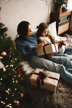 Happy woman laying in bed with kid girl open Christmas present boxes in bedroom. Winter holiday season. Cozy home atmosphere.