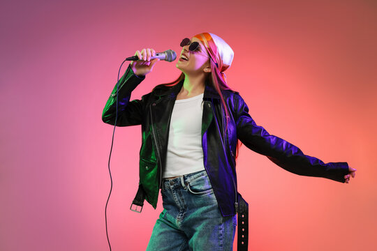 Smiling singer in bandana, sunglasses and leather jacket with microphone performing in color lights on pink gradient background