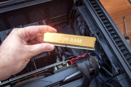 Gold DDR RAM module in a technician's hand. Concept of rising RAM prices. Expensive computer parts and difficult upgrades. Open desktop PC case with powerful graphics card and cooling fan