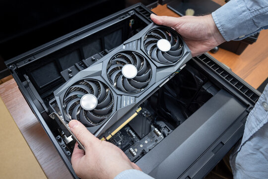 Technician holds a powerful expensive gaming graphics card with three fans above an open desktop PC case. Assembly or upgrade of a powerful gaming PC