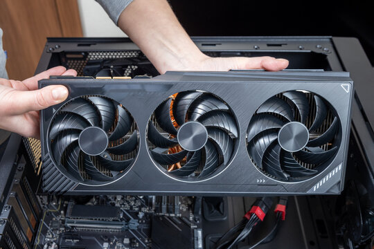 A technician installs a powerful triple-fan gaming graphics card on a gaming PC motherboard. A dual-tower CPU cooler and black power cables are visible