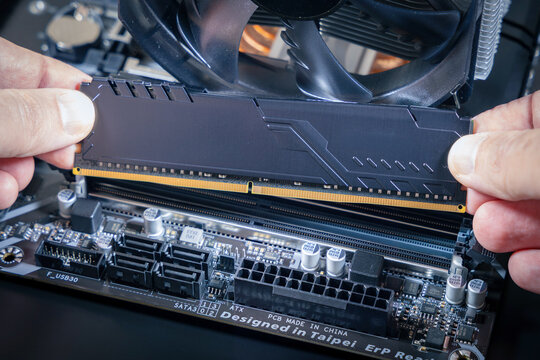 Hands hold a DDR memory module above a DIMM slot on a motherboard. SATA ports, an ATX power connector, and a CPU fan are visible. Modern PC assembly or upgrade