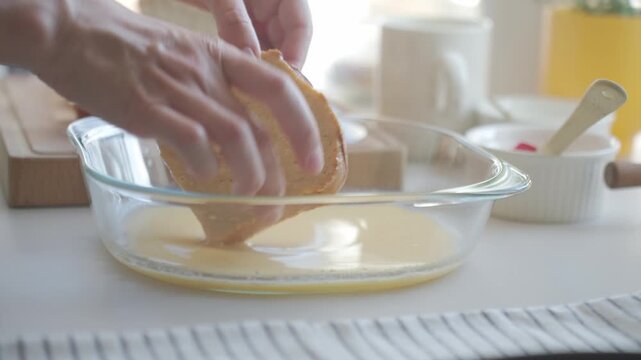 Hand dips bread in egg mixture in glass dish on kitchen counter