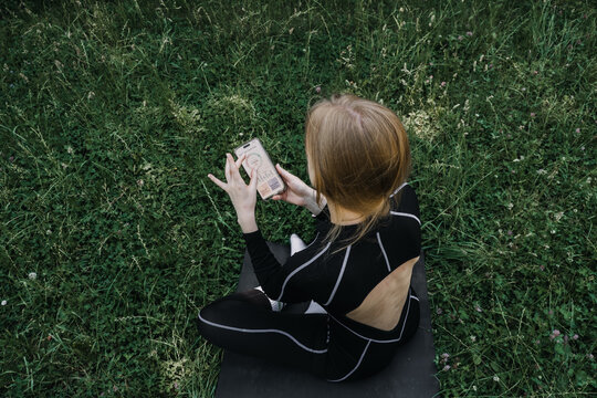 Woman sits on grass holding smartphone displaying hormone tracking application. cycle syncing workout, menstrual cycle app, femtech tracking, women health technology