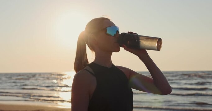 sporty woman drinking water from bottle on beach after outdoor workout