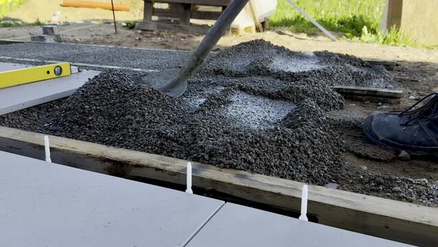 Worker spreads wet concrete with a shovel on a floor surface, with tools and wooden supports visible in the outdoor construction area