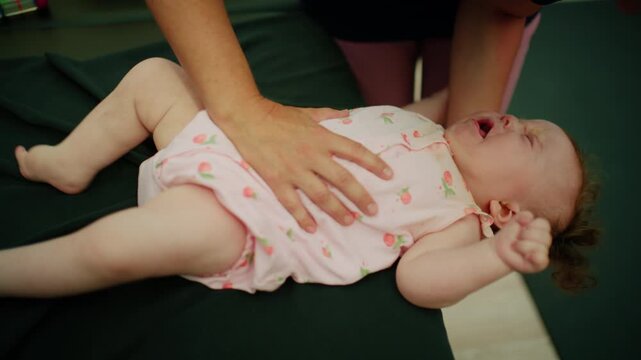 Baby crying on a examination table while a therapist is performing a gentle massage on her abdomen for colic relief