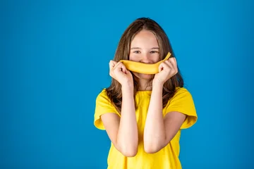 Playful young girl holding a banana in front of her mouth like a smile, wearing a yellow t-shirt against a blue studio background © Nikolay N. Antonov