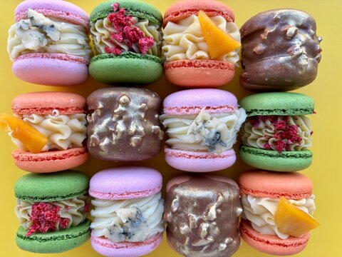 Flat lay of a 3x4 grid showcasing colorful gourmet macarons with artisanal fillings on a bright yellow background. Features unique flavors including macarons stuffed with blue cheese, dried raspberrie