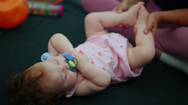 Baby girl sleeping calmly with a pacifier, wearing a pink romper, while receiving caress on her leg representing care and parenthood