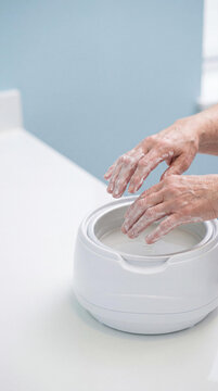 Elderly woman applying paraffin wax bath to hand. Senior patient using thermal therapy for joint pain relief and arthritis treatment. Medical care concept for hand health and wellness.