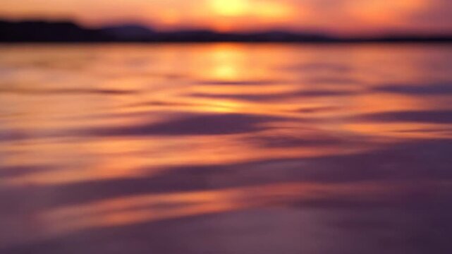 Abstract blurred water ripples reflecting golden sunset light. Soft bokeh effect on calm surface with vibrant orange and purple tones and distant horizon.