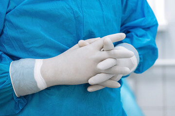 Fototapeta premium Medical professional wearing blue scrubs and white gloves stands with hands clasped in a clinical setting, emphasizing hygiene and readiness for patient care