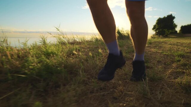 A person is jogging along a grassy path by the water at sunset. The runner's legs and feet are visible, showcasing a healthy lifestyle through this sport activity.