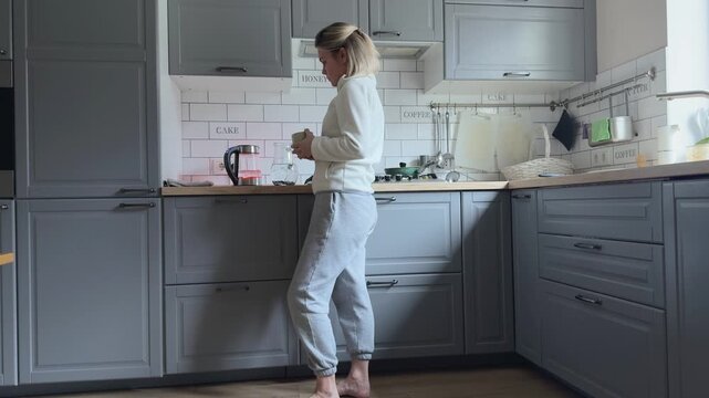 Woman in light home clothes stands by the countertop waiting for the electric kettle to boil in a gray kitchen with white tile backsplash, soft lighting and a clean minimalist look.