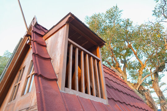 A rustic wooden dormer window on a red tiled roof with leafy trees in the background.