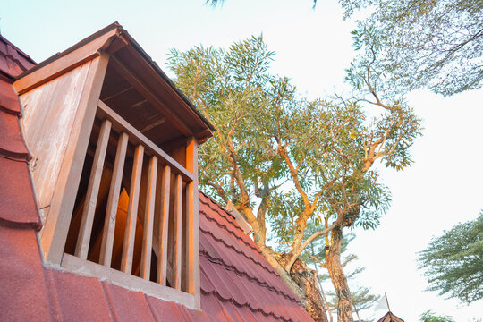 A rustic wooden dormer window on a red tiled roof with leafy trees in the background.