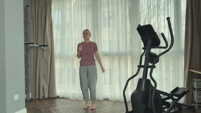 Young woman performs light bouncing exercise on straight legs in a bright modern living room, stimulating lymphatic drainage and circulation. She is dressed in a soft pink short sleeve t shirt and