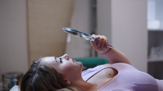 Woman checking new eyelash extensions in a hand mirror at a clinic