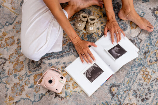 Top down view of a pregnant woman sitting on a rug looking at an open pregnancy album with an ultrasound photo an instant camera and baby booties a cozy maternity scene.