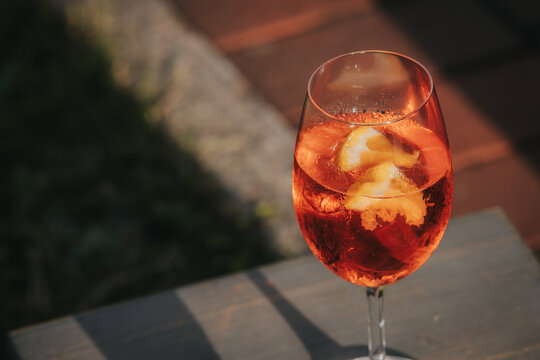 Glass of orange cocktail with ice cubes and lemon slices placed on a wooden table, outdoor setting with blurred background of grass and stone pathway