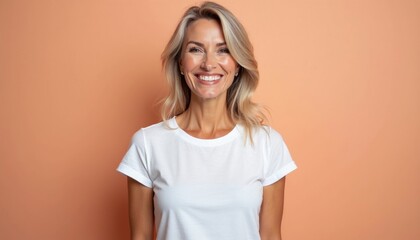 Obraz premium Mature woman with blonde hair smiles wearing white t-shirt against peach background. Face shows natural makeup, healthy skin. Looks directly at camera with friendly expression. Portrait highlights