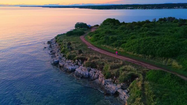 A senior woman jogs along a scenic coastal path surrounded by lush greenery and tranquil waters. The wide shot captures her solitary run at sunrise, highlighting her dedication to a healthy lifestyle.