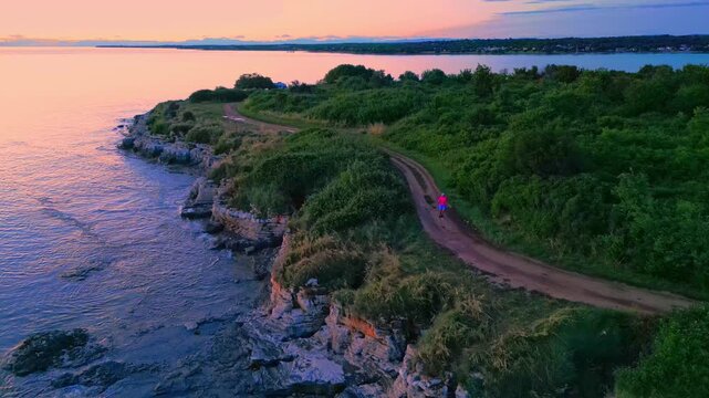 A person jogs along a serene coastal path at sunset, surrounded by lush greenery and the calm sea, highlighting nature's tranquility and dedication to a healthy lifestyle.