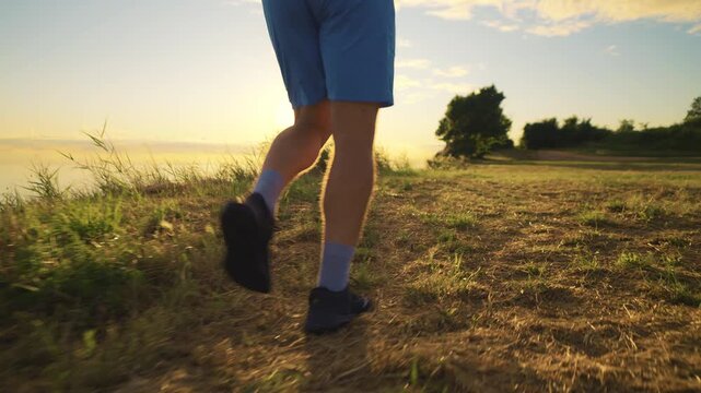 A senior adult jogs along a sunlit path, wearing blue shorts and black shoes. The camera captures their determined strides from a low angle, emphasizing movement against the serene, grassy landscape.