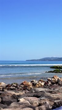 Volcanic rocky shoreline, tourist destination gentle waves breaking under clear midday sky, wet barnacled boulders, green algae, reflective surf, distant forested peninsula and calm coastal horizon
