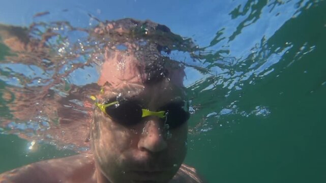 Man swimming with goggles, creating surface water reflection and distortions from an underwater perspective, with bright sky above