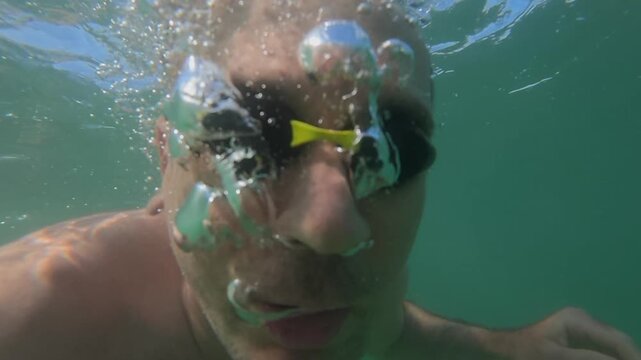 Person wearing swimming goggles with face partially submerged, looking up from below the surface with water bubbles