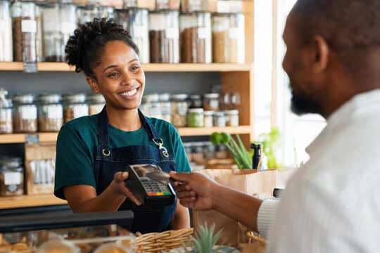 Male customer buys fresh, healthy groceries and pays with debit card. At counter with different products, African American merchant holds POS for black woman to make payment.
